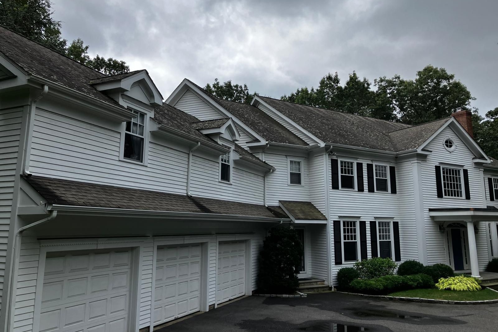 Clapboard Siding Installation & Refinishing – A view of a house with white clapboard siding, showcasing a well-maintained exterior with black shutters, garage doors, and lush landscaping.